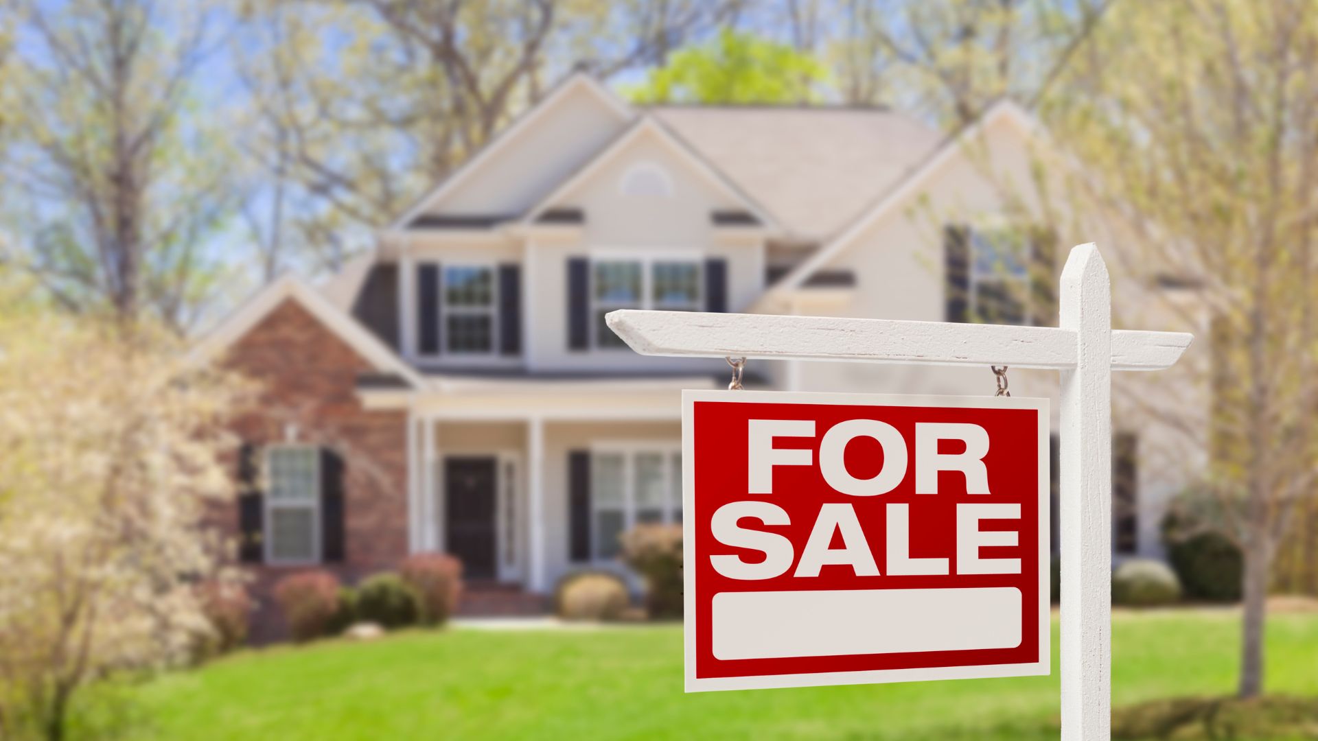 A house with a "For Sale" sign in the foreground against a backdrop of trees and a clear sky.