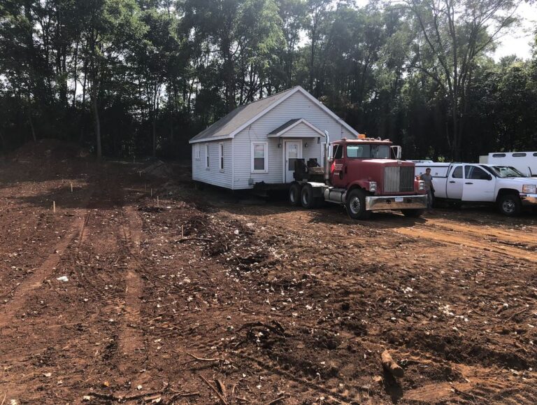 A house is being relocated by a red truck on a dirt lot surrounded by trees, with additional white vehicles nearby, likely from a tree company assisting the move.