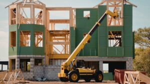 structural lifting in michigan yellow telescopic forklift lifts materials to the upper floor of a partially constructed two-story house with green sheathing and exposed wooden framing.