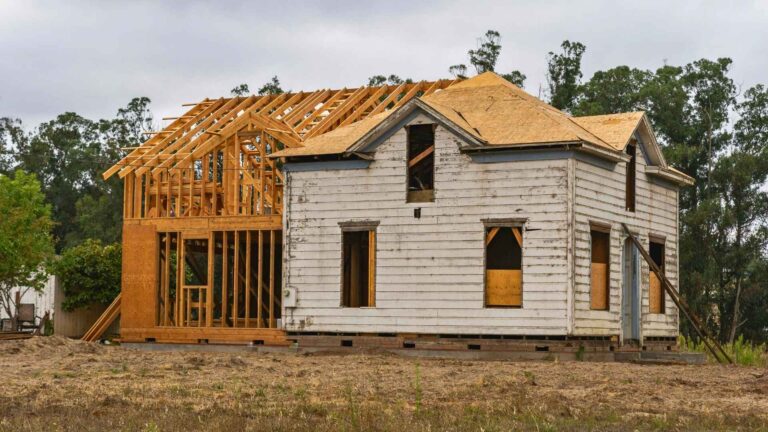 structure moving michigan A partially renovated house with an old painted exterior and new wooden framing being added to one side, set on a dirt lot with trees in the background.